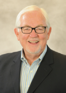 Albert M. (Al) Erisman with glasses, in dark blazer and striped shirt, smiles in front of a light indoor background.